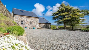 A stone house with a sloped roof, a gravel driveway, and a large tree in the foreground. - Two bedroom barn conversion on Anglesey | Bwthyn-Cerrig (Llannerch-y-medd)
