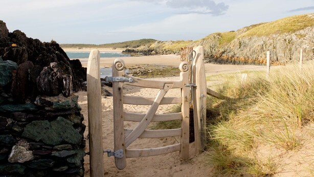 A wooden gate leading to a sandy beach with a stone wall on one side and grassy dunes on the other. - Two bedroom barn conversion on Anglesey | Bwthyn-Cerrig (Llannerch-y-medd)