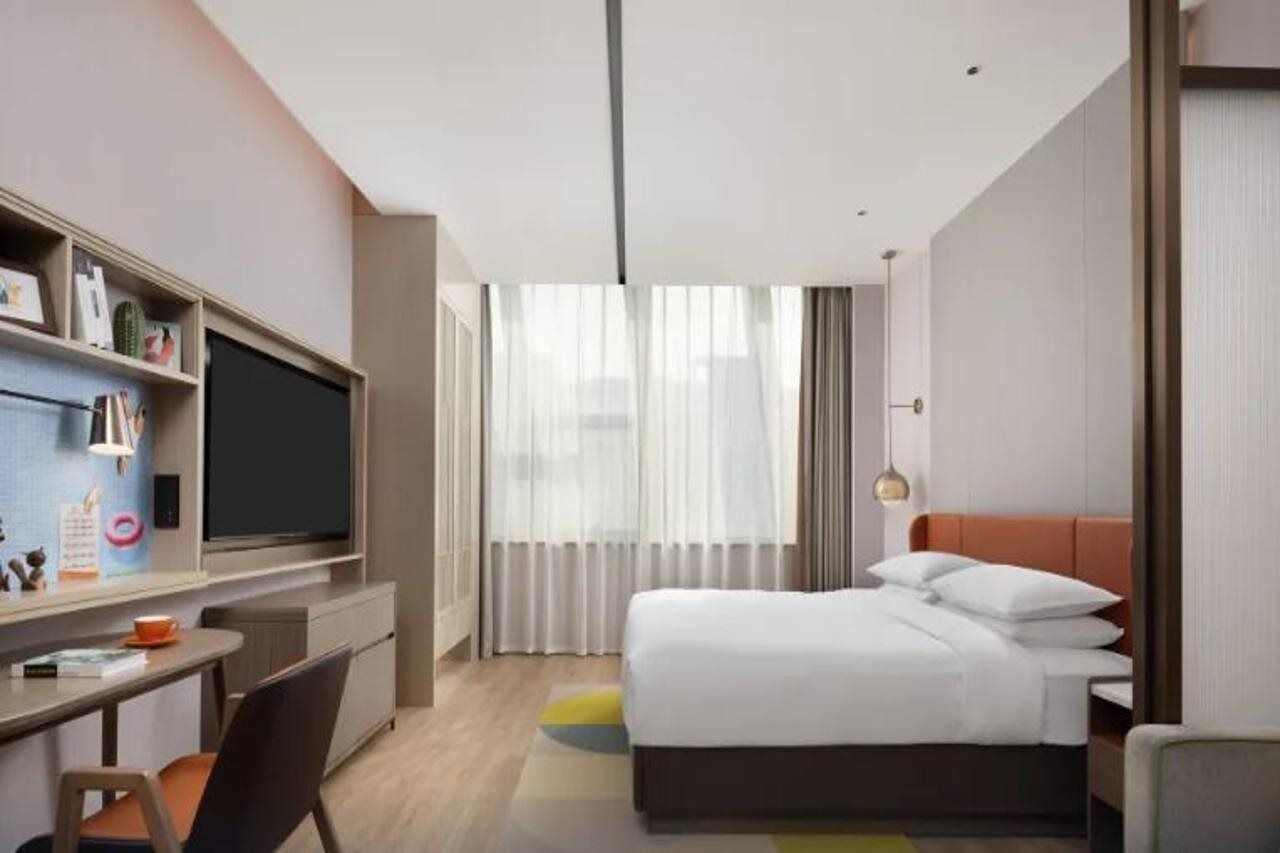 A modern hotel room with a bed, a desk with a computer, a television, and a window with curtains.