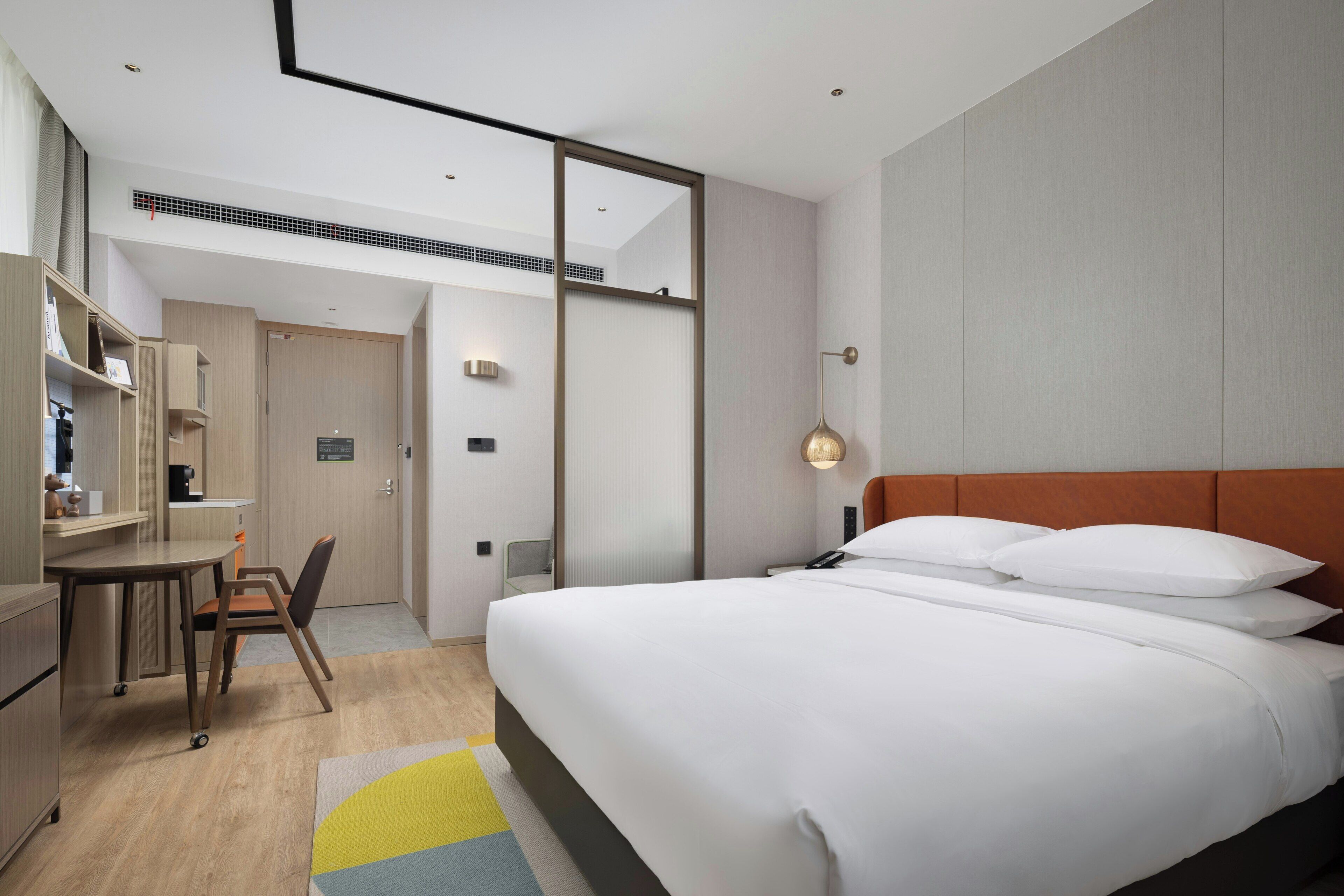 A hotel room with a bed, desk, chair, and a view of the hallway.