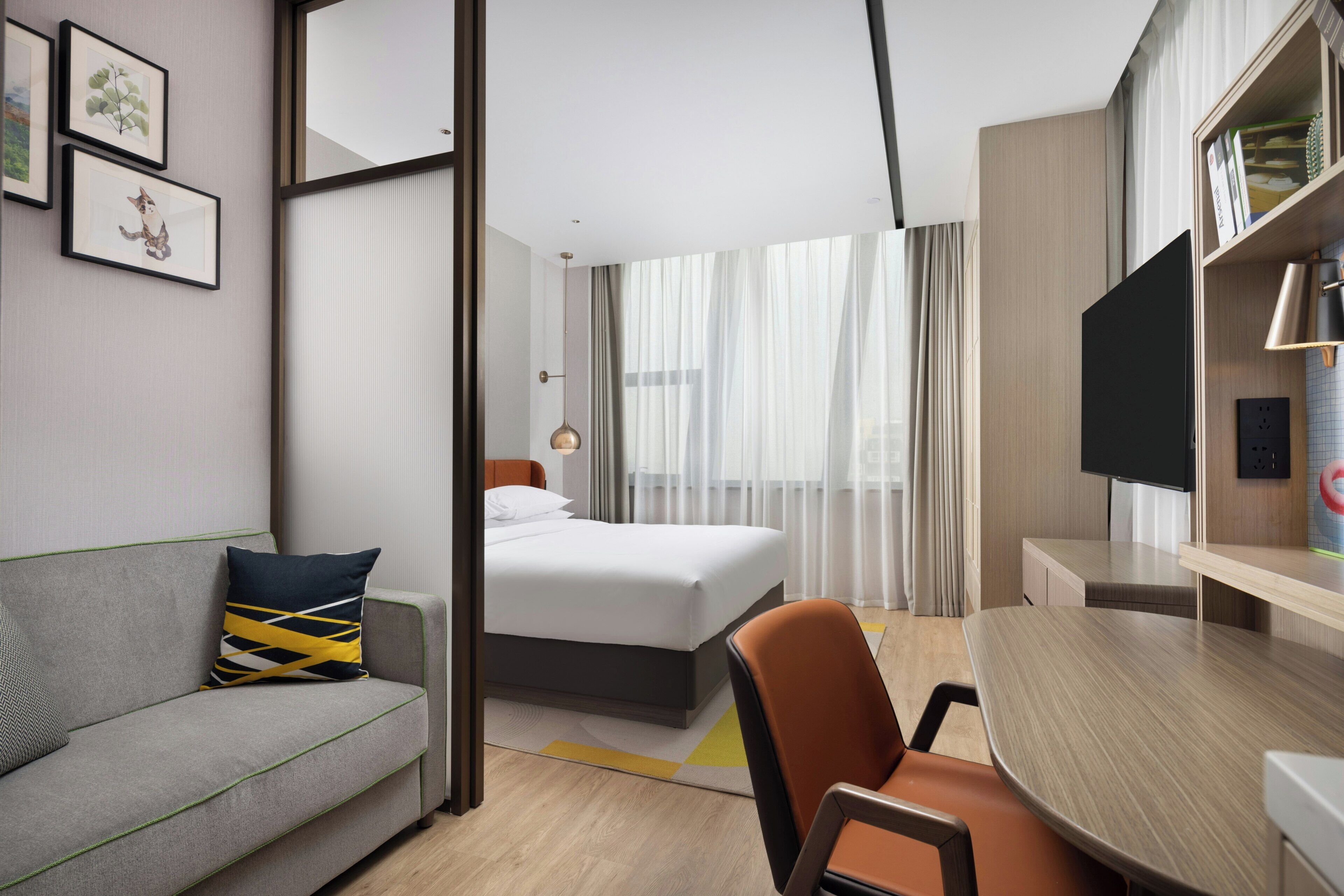 A hotel room with a bed, a sofa, a desk, a chair, a TV, and a bookshelf.