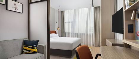 A hotel room with a bed, a sofa, a desk, a chair, a TV, and a bookshelf.