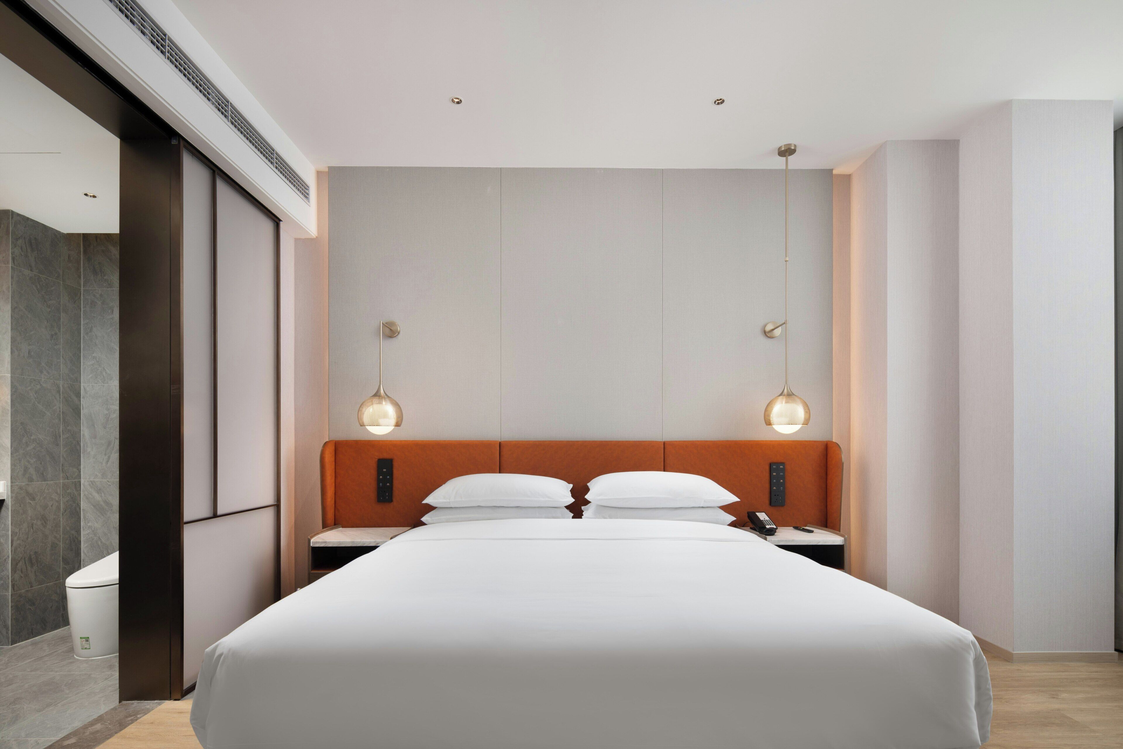A modern hotel room with a large bed, two bedside tables, a nightstand, a wall-mounted control panel, and a large window.