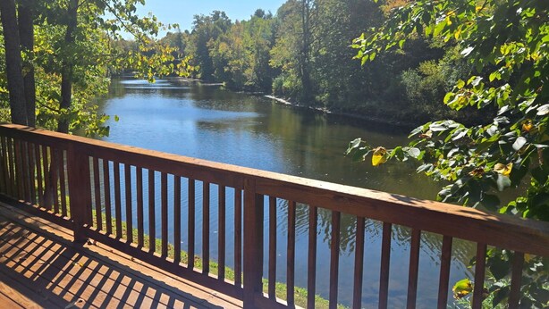A wooden deck with a railing overlooking a calm river surrounded by trees. - Cozy cabin in Brasher Falls, NY (Brasher Falls)
