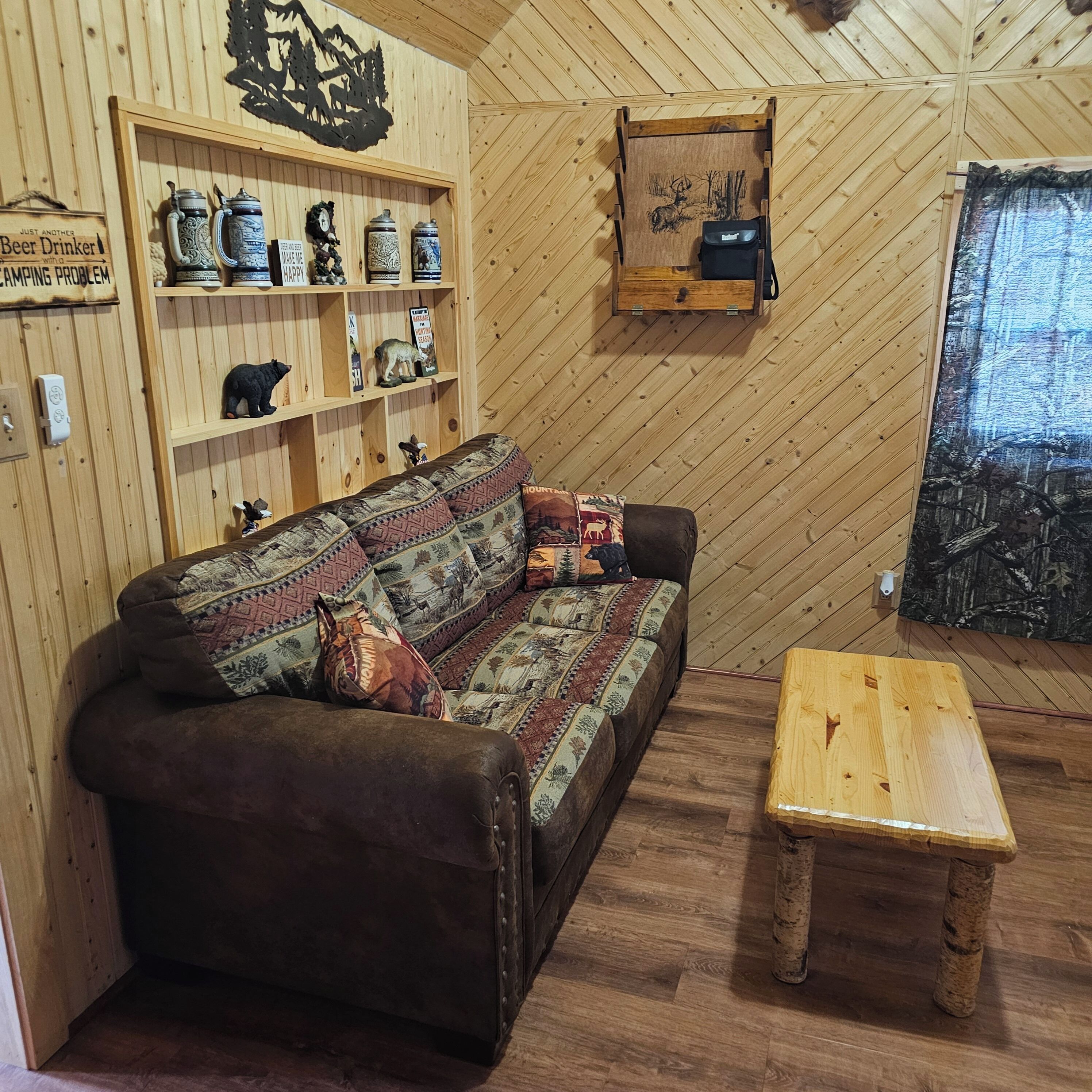 Una habitación acogedora con suelo de madera en espiga, un sofá marrón con cojines estampados, una pequeña mesa de madera y un estante fijado a la pared con diversos objetos.