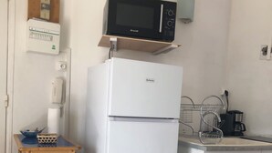 Una cocina compacta con un refrigerador blanco, un microondas encima, una mesita de madera y una estantería de metal.