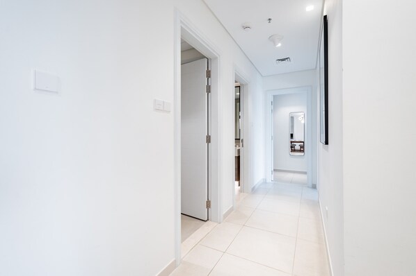 A modern, well-lit hallway with white walls and a tiled floor, leading to a room with a door.