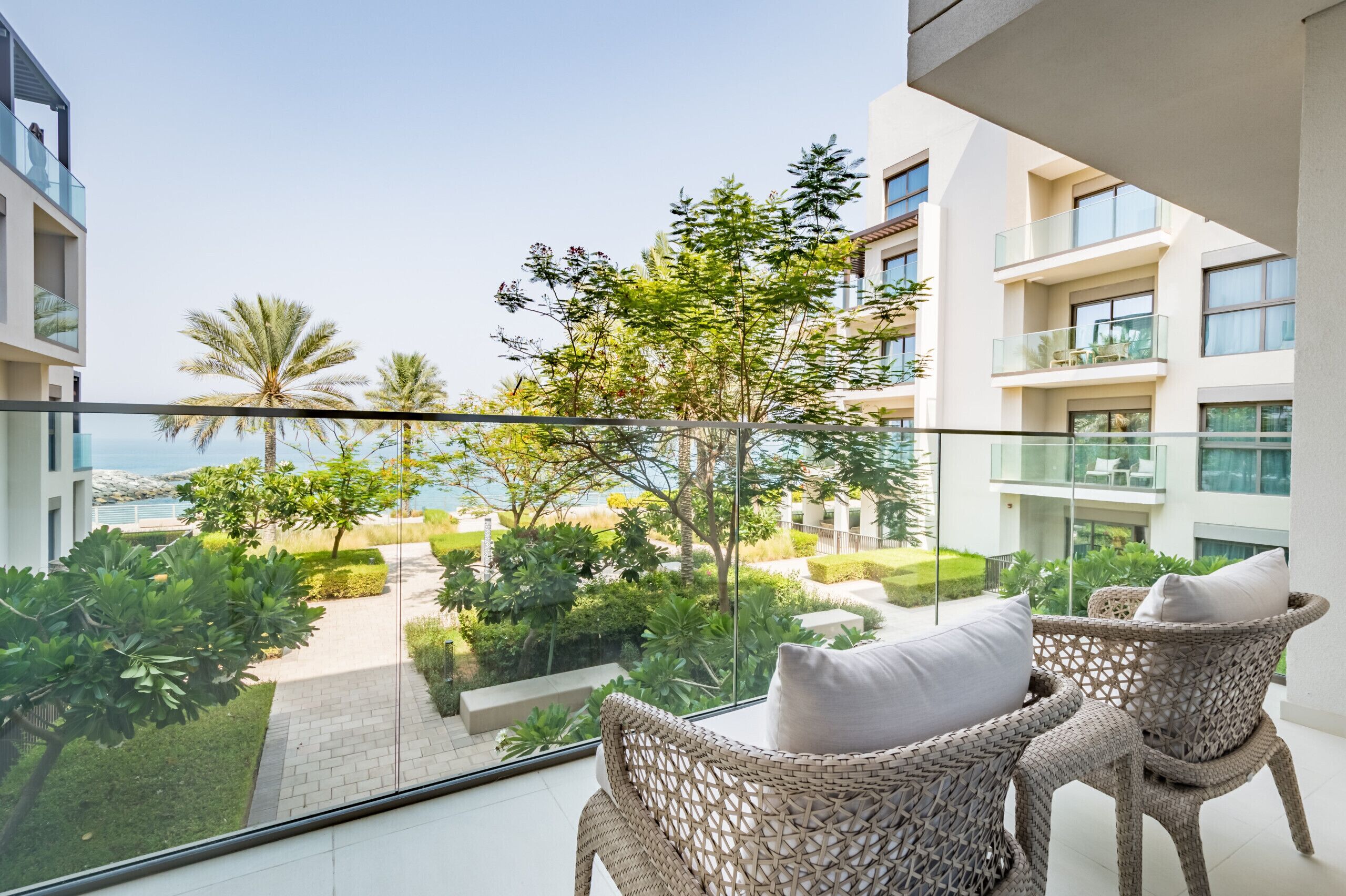 Ein Balkon mit Korbstühlen, einer Glasreling und Blick auf einen angelegten Garten sowie Gebäude.