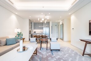 A modern living room with a sofa, coffee table, and a dining area with chairs. There is a kitchen in the background with cabinets and a refrigerator.