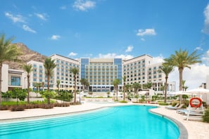 A hotel with a large swimming pool, palm trees, and a mountainous backdrop. - Waterfront 2BR Apartment in The Address Fujairah Residences Block C, Fujairah (Fujairah)