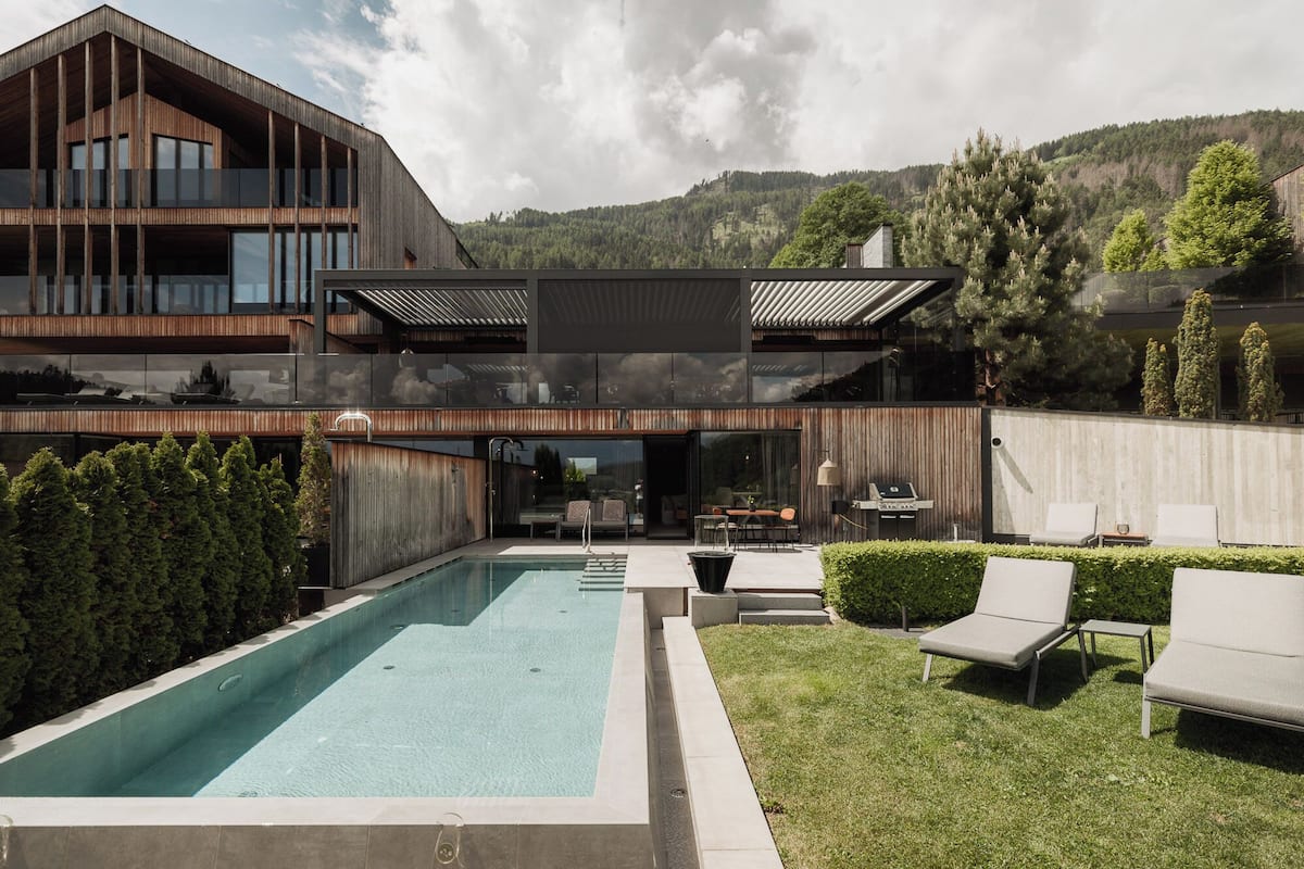 A modern house with a large swimming pool, outdoor seating, and a view of a forested hillside.