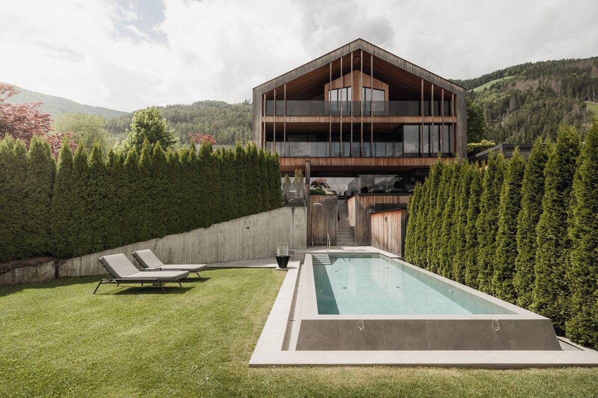 A modern two-story house with a swimming pool, surrounded by a well-maintained lawn and dense hedges.