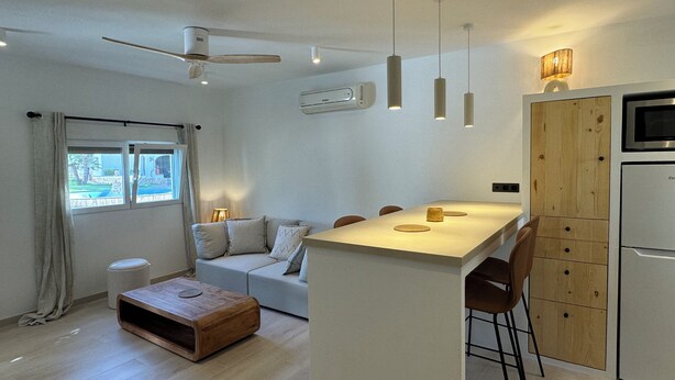Een moderne keuken met een centraal eiland, een eetgedeelte met stoelen, een bank en een houten salontafel. Er is een raam met gordijnen, een plafondventilator en hanglampen.