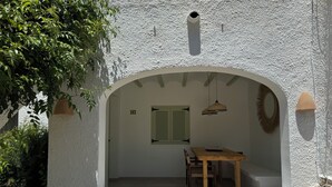 Een kamer met witte wanden, een houten tafel en stoelen, een raam met luiken en het nummer '103' op de muur.