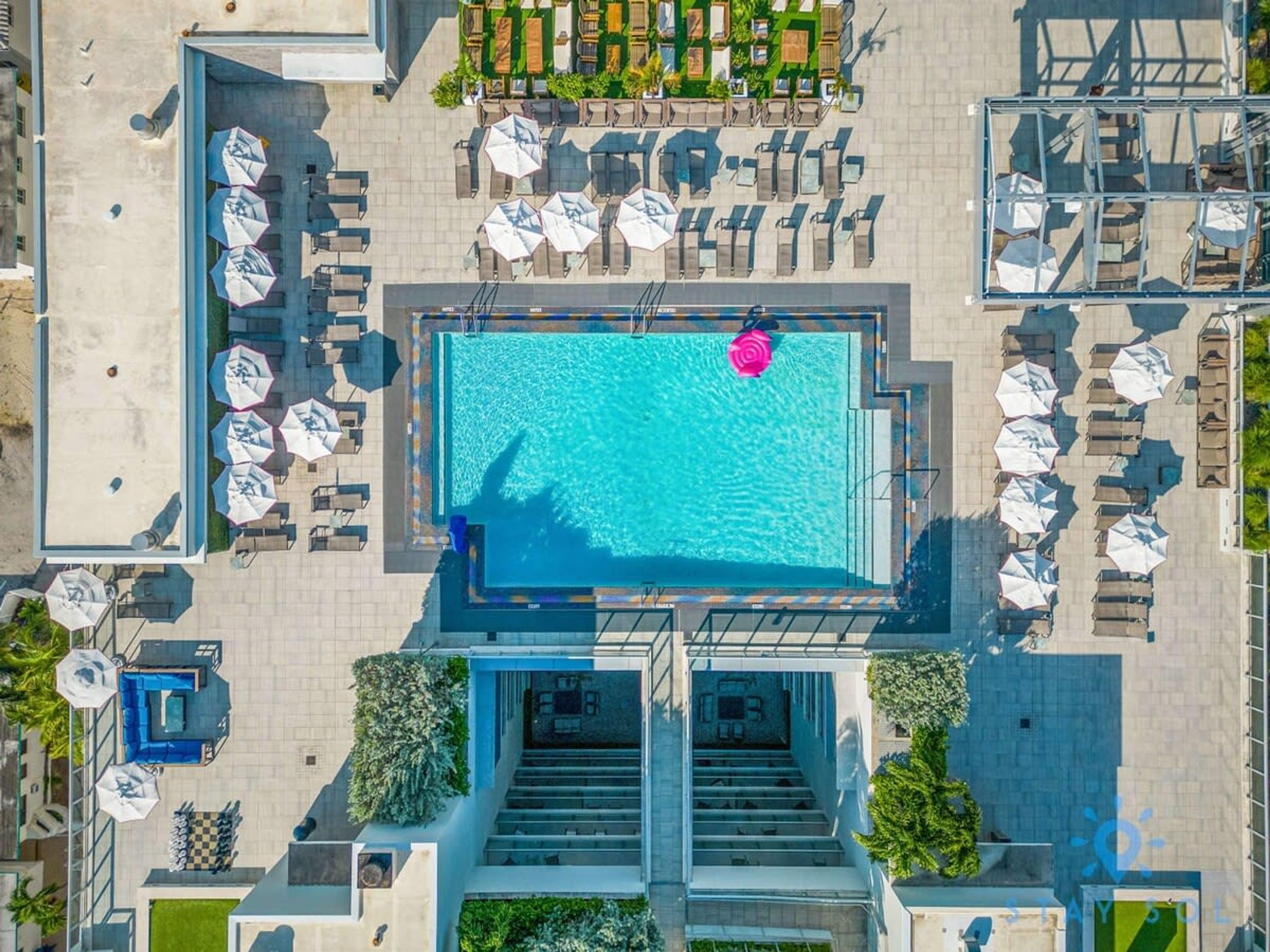 A rooftop pool area with multiple lounge chairs and umbrellas, surrounded by a building with a tiled floor.