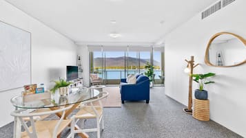A modern living room with a glass table, chairs, a blue sofa, a large mirror, and a view of a mountainous landscape through large windows.