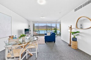 Un salon moderne avec une table en verre, des chaises, un canapé bleu, un grand miroir et une vue sur un paysage montagneux par de grandes fenêtres.