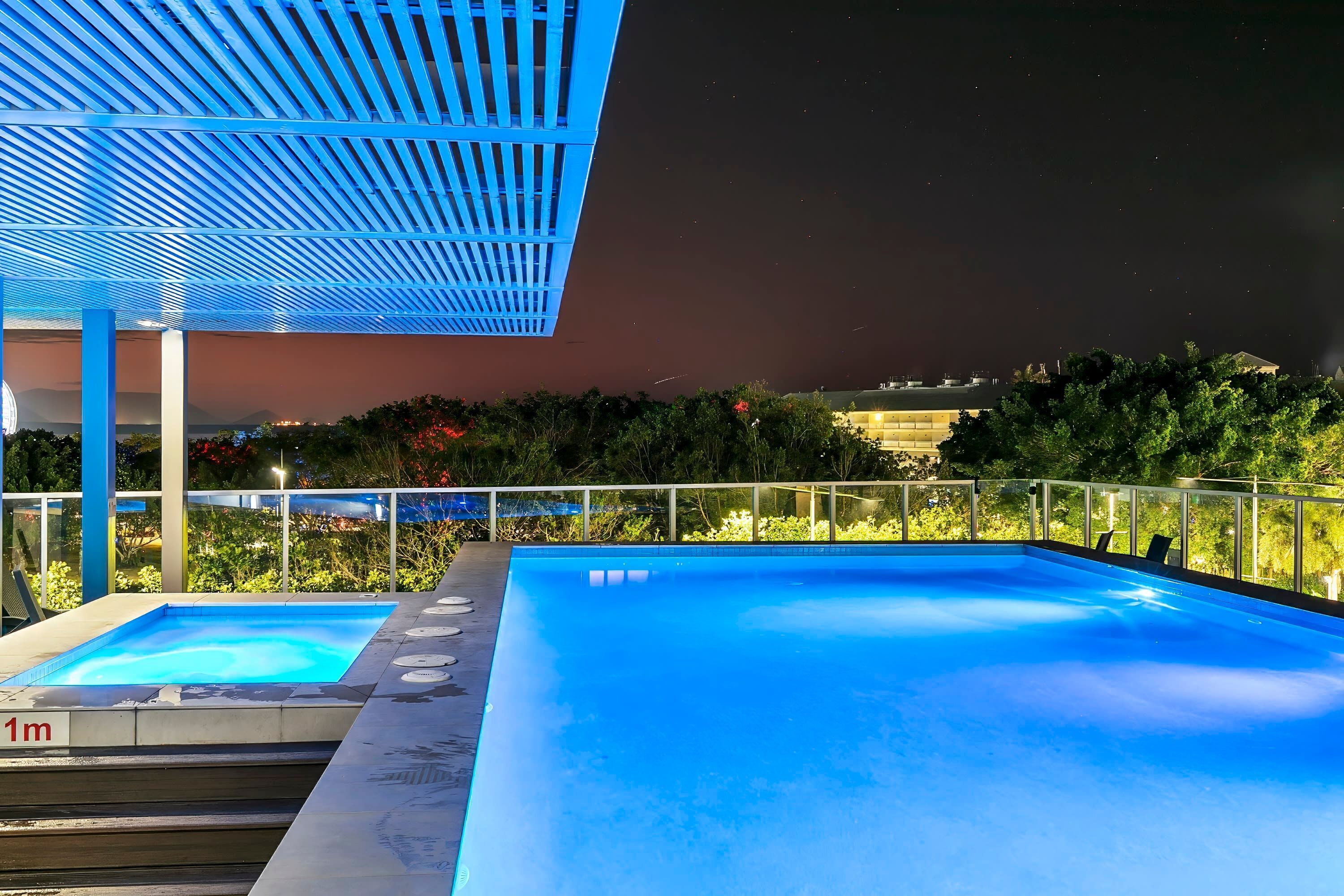 Piscina no terraço iluminada em azul, com toldo, cercada por vegetação e guarda-corpo de vidro.