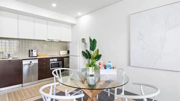 Una cocina moderna con armarios blancos, un lavavajillas de acero inoxidable y un microondas. Hay una mesa de vidrio con sillas blancas y una planta en maceta.