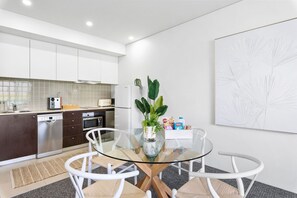 Une cuisine moderne avec des meubles blancs, un lave-vaisselle en acier inoxydable et un four à micro-ondes. On y trouve une table en verre, des chaises blanches et une plante en pot.