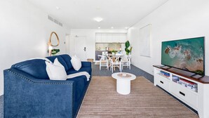A modern living room with a blue sofa, a coffee table, and a TV on a white stand. - Lux Waterfront - Prime Location Pool Gym Sauna (Cairns)