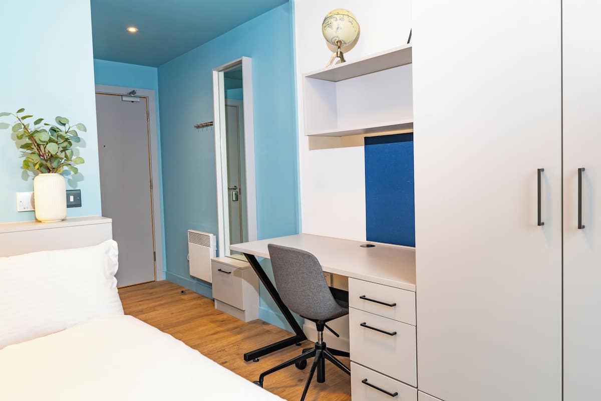 A bedroom with a bed, a desk, a chair, a plant, and a wardrobe.