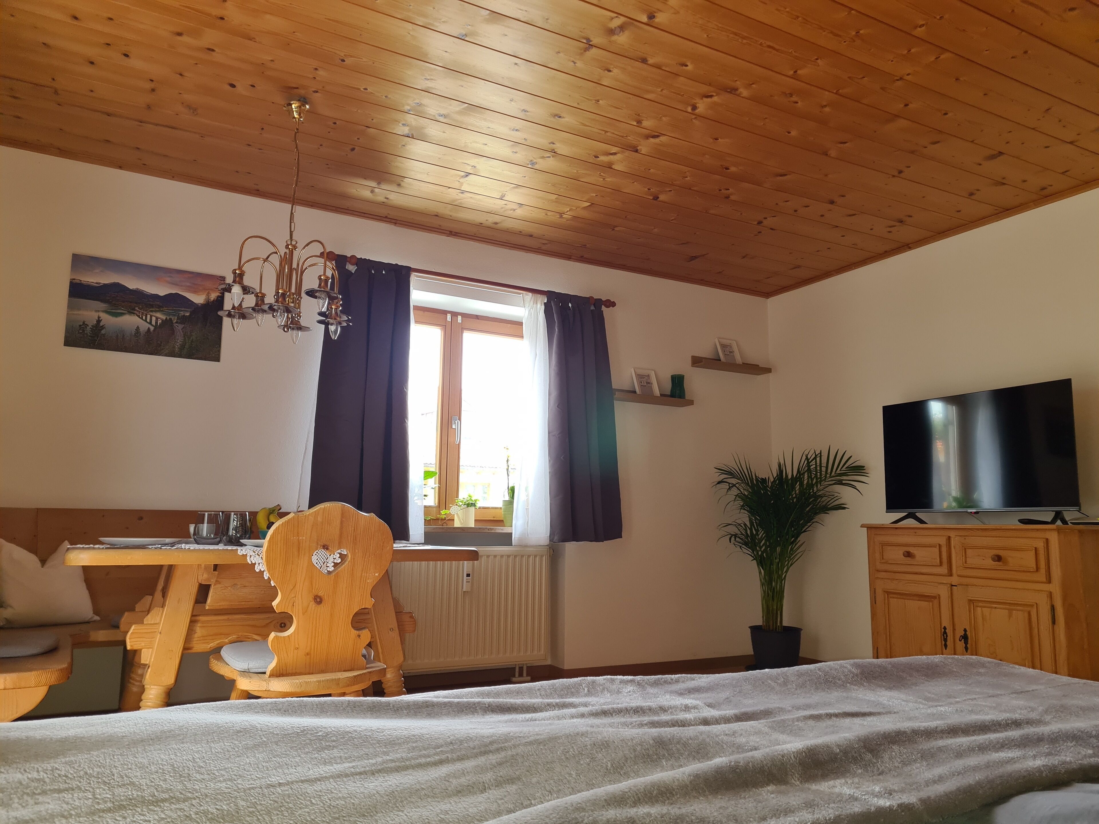 Ein gemütliches Zimmer mit Holzbalkendecke, einem Esstisch mit Stühlen, einem Bett mit weißer Decke, einem Fernseher auf einer Holzkommode und einem Fenster mit Vorhängen.