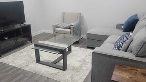 A modern living room with a flat-screen TV, a glass coffee table, a grey sofa, and a beige armchair. - A Cozy home away from home at South East Calgary (Calgary)