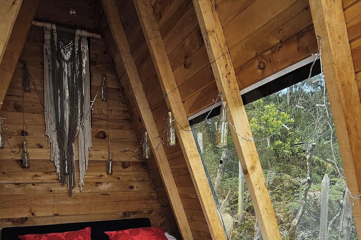 Une chambre à coucher dans une cabane en bois, avec un plafond en pente, un lit agrémenté d’oreillers rouges et une vue sur la forêt par la fenêtre.