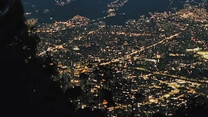 Paisatge urbà nocturn amb una muntanya al primer pla i una zona urbana il·luminada al fons.