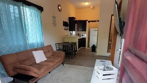 Een gezellige woonkamer met een bruine bank, een witte bijzettafel en een eethoek, met een keuken op de achtergrond.