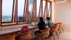 Une pièce avec une longue table et plusieurs chaises en bois, de grandes fenêtres donnant sur un paysage verdoyant et des bâtiments, et un climatiseur fixé au mur.