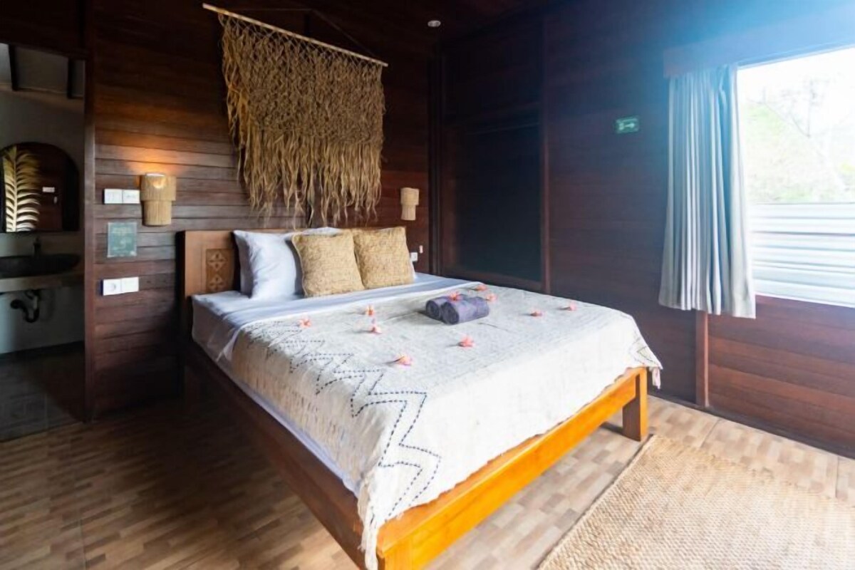 A wooden-framed bed with a white bedspread, a hanging macrame piece, and a window with curtains.