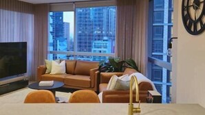 Una sala de estar moderna con un sofá, una mesa de centro y un televisor. Se ve una ventana grande con vistas a edificios.