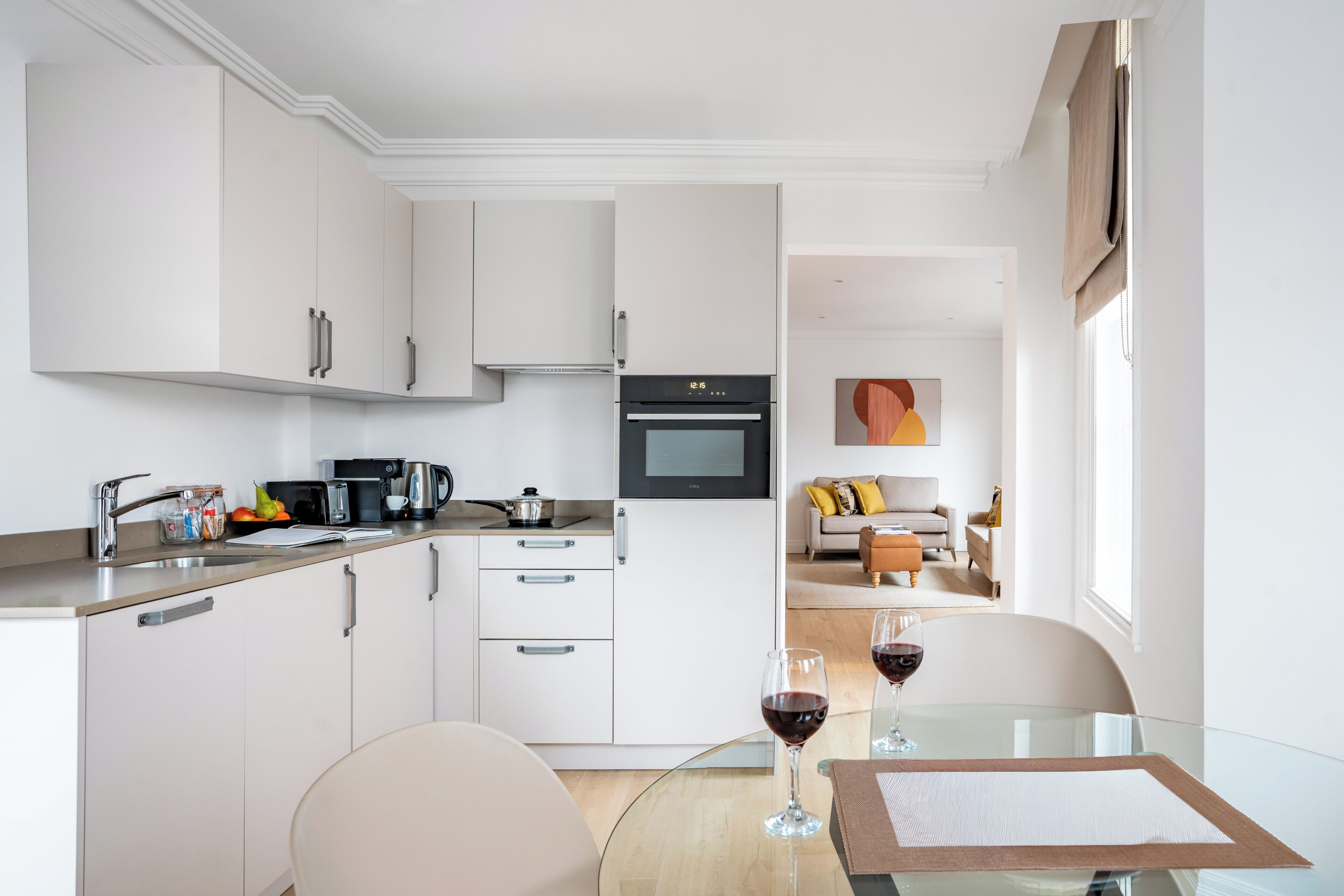 Una cocina moderna con armarios blancos, horno empotrado y un comedor con mesa y sillas de vidrio.