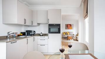 Una cocina moderna con armarios blancos, horno empotrado y un comedor con mesa y sillas de vidrio.