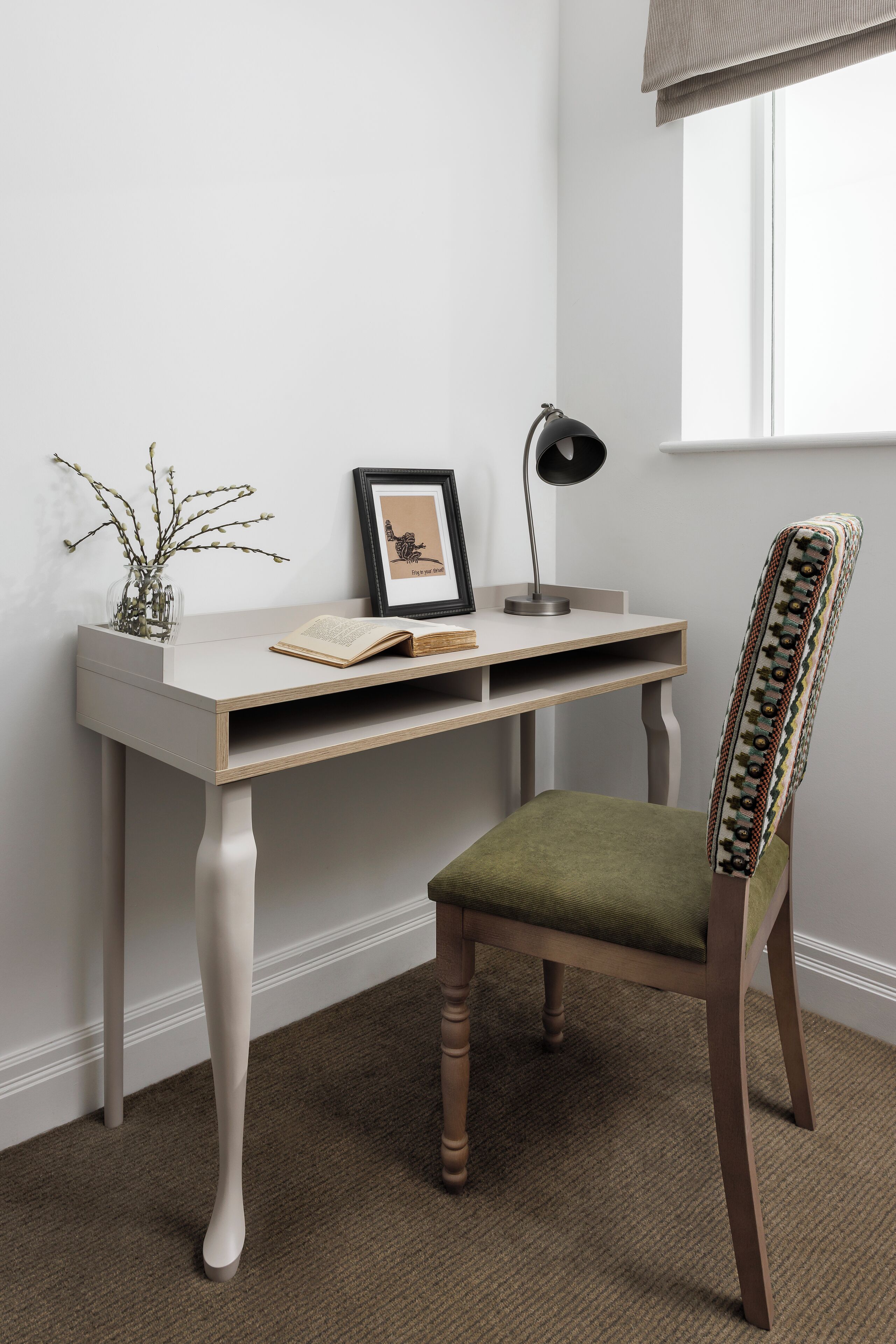Un espace de travail comprenant un bureau, une chaise, une lampe, un tableau encadré et un vase avec des branches.