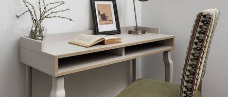 A study area with a desk, chair, lamp, framed picture, and a vase with branches.