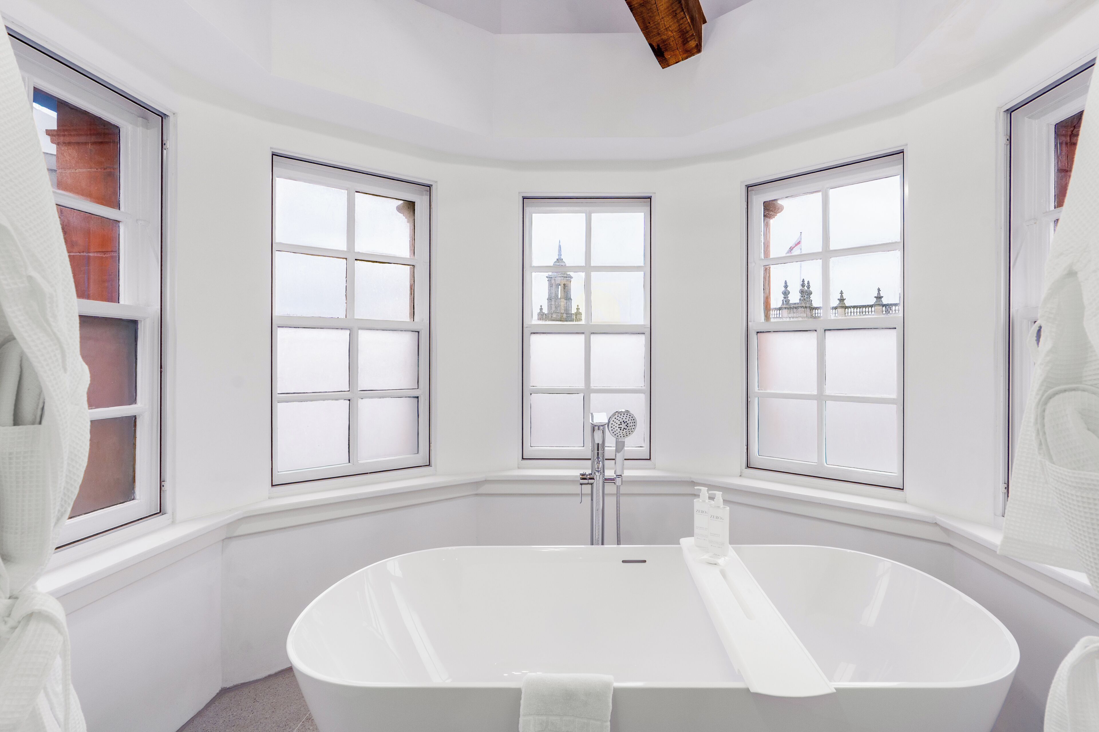 A corner bathroom with a freestanding bathtub, three windows, and a view of a distant building.