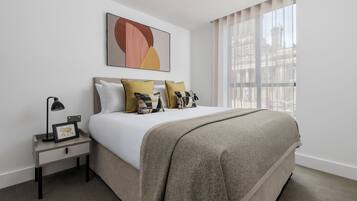 Ein ordentlich eingerichtetes Hotelzimmer mit Bett, Nachttisch mit Lampe und großem Fenster mit Vorhängen.