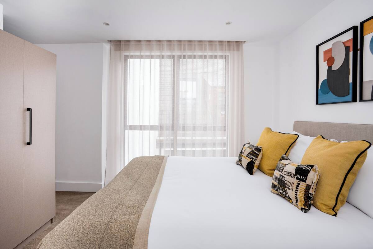A hotel room with a bed, a wardrobe, a window with sheer curtains, and a framed abstract painting on the wall.