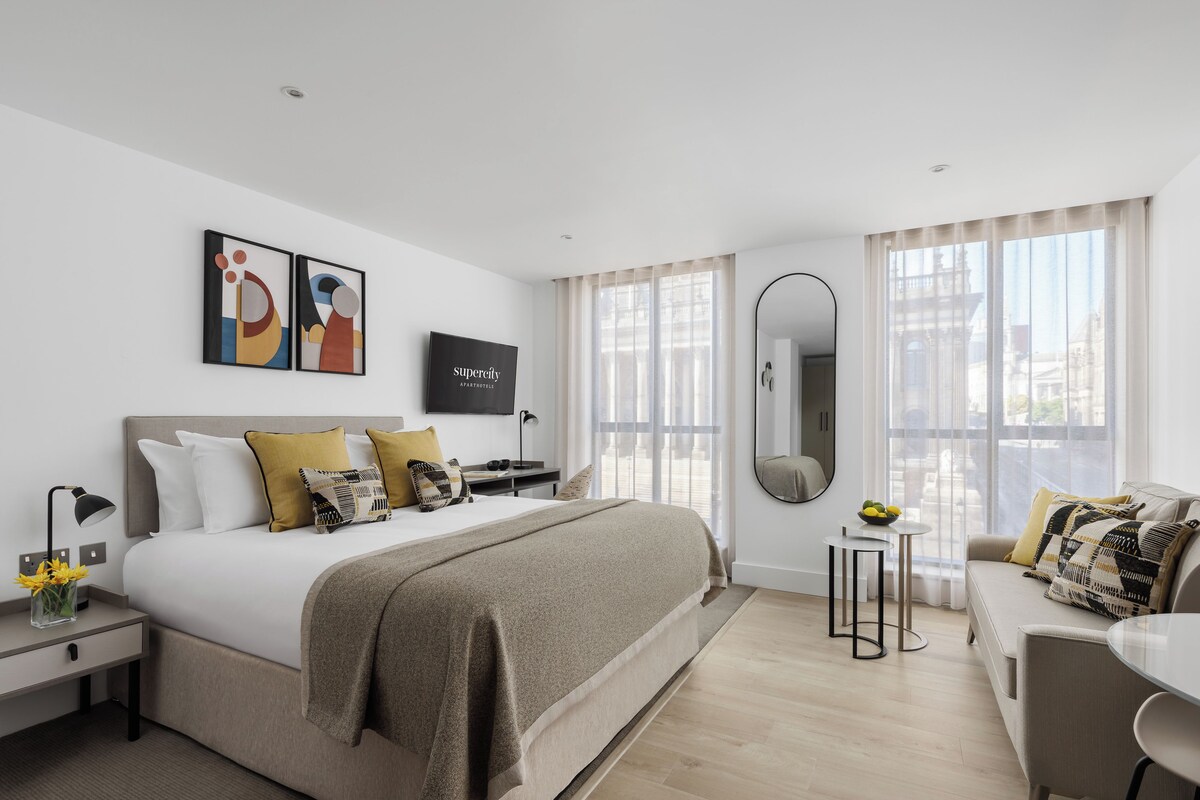 A modern hotel room with a large bed, a desk, a chair, a TV, and a view of the city through the window.