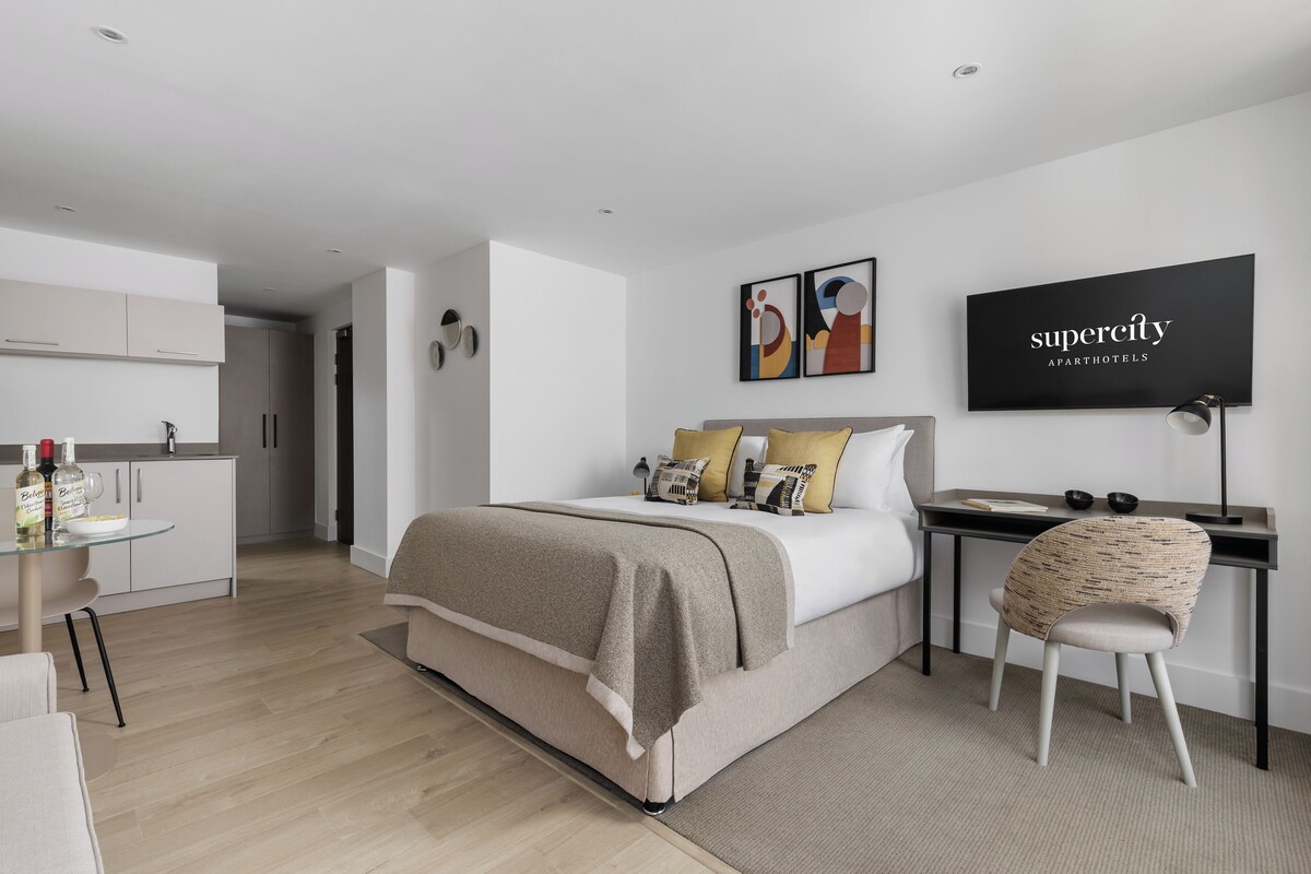 Un dormitorio moderno con cama, mesita de noche y escritorio con silla. Se observa una cocina al fondo.