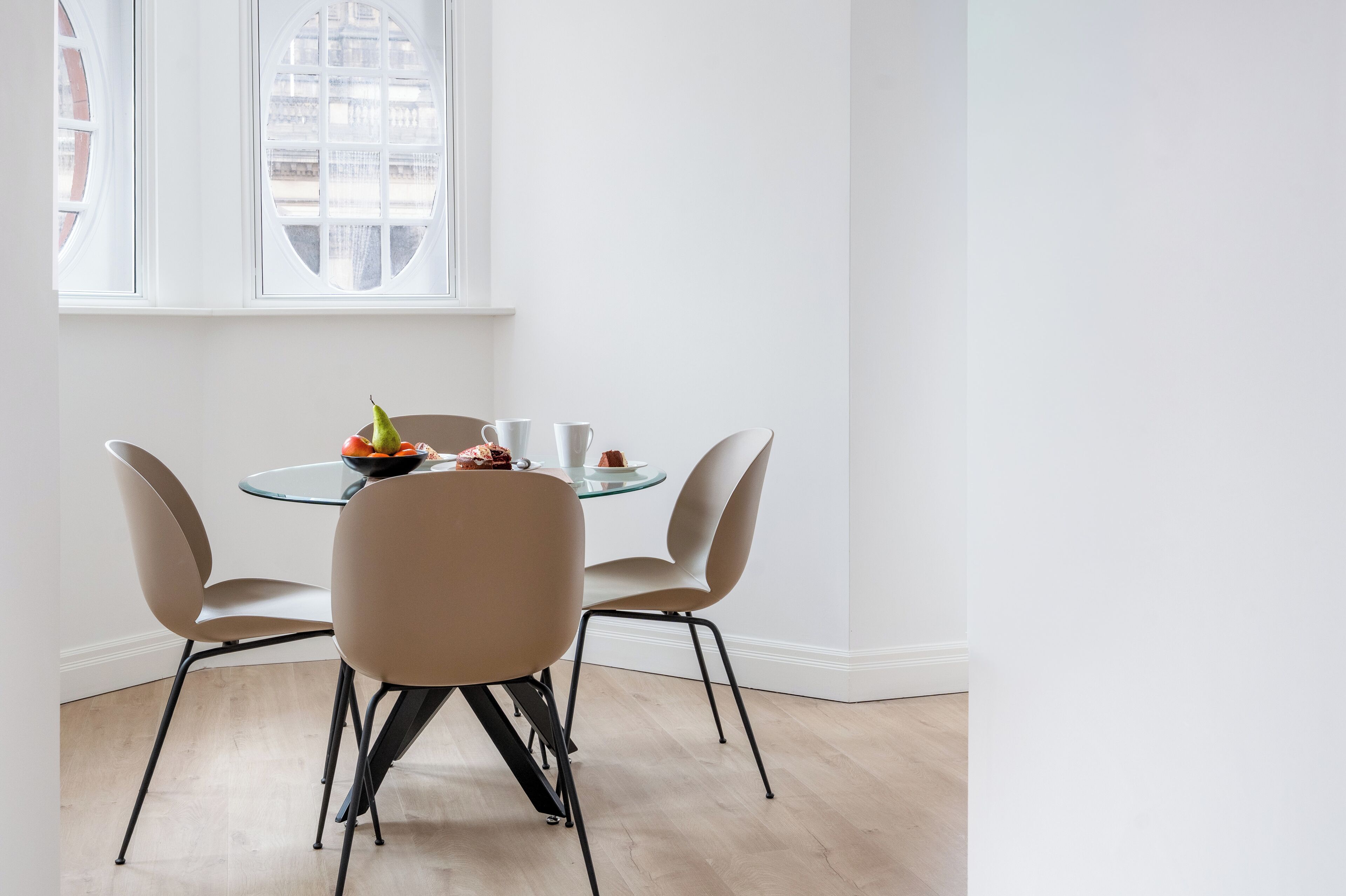 A round glass table with four chairs, a bowl of fruit, and two cups on the table.
