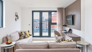 A modern hotel room with a sofa, a desk with a laptop, a TV, and a large window with a city view.