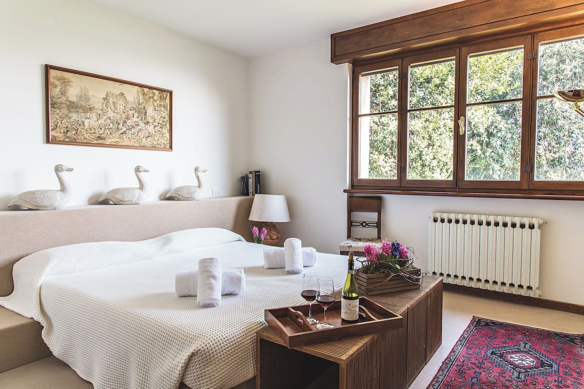 Ein Hotelzimmer mit einem Bett, einem Nachttisch aus Holz mit einer Weinflasche und Gläsern, einem Heizkörper und einem Fenster mit Blick auf Bäume.
