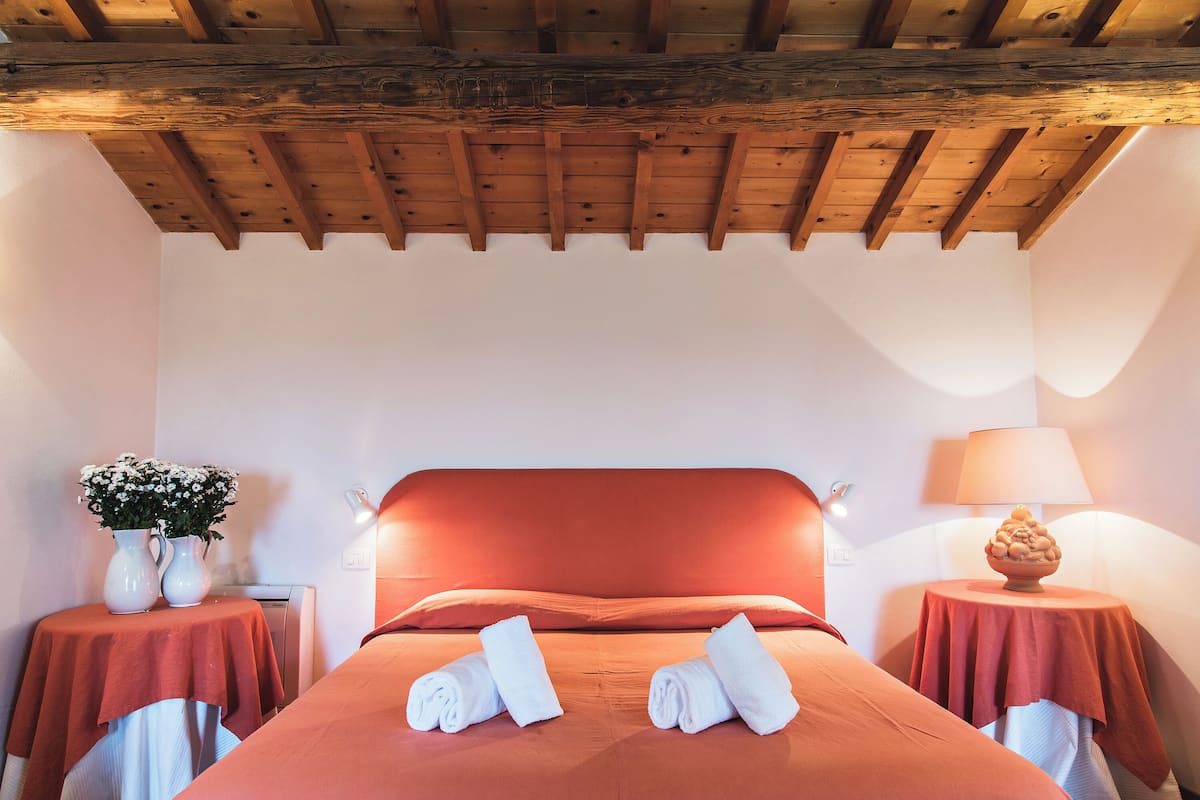 Ein Hotelzimmer mit Holzbalkendecke, einem Bett mit roter Bettwäsche, zwei Nachttischen mit Lampen und einem Blumenstrauß auf einem Tisch.