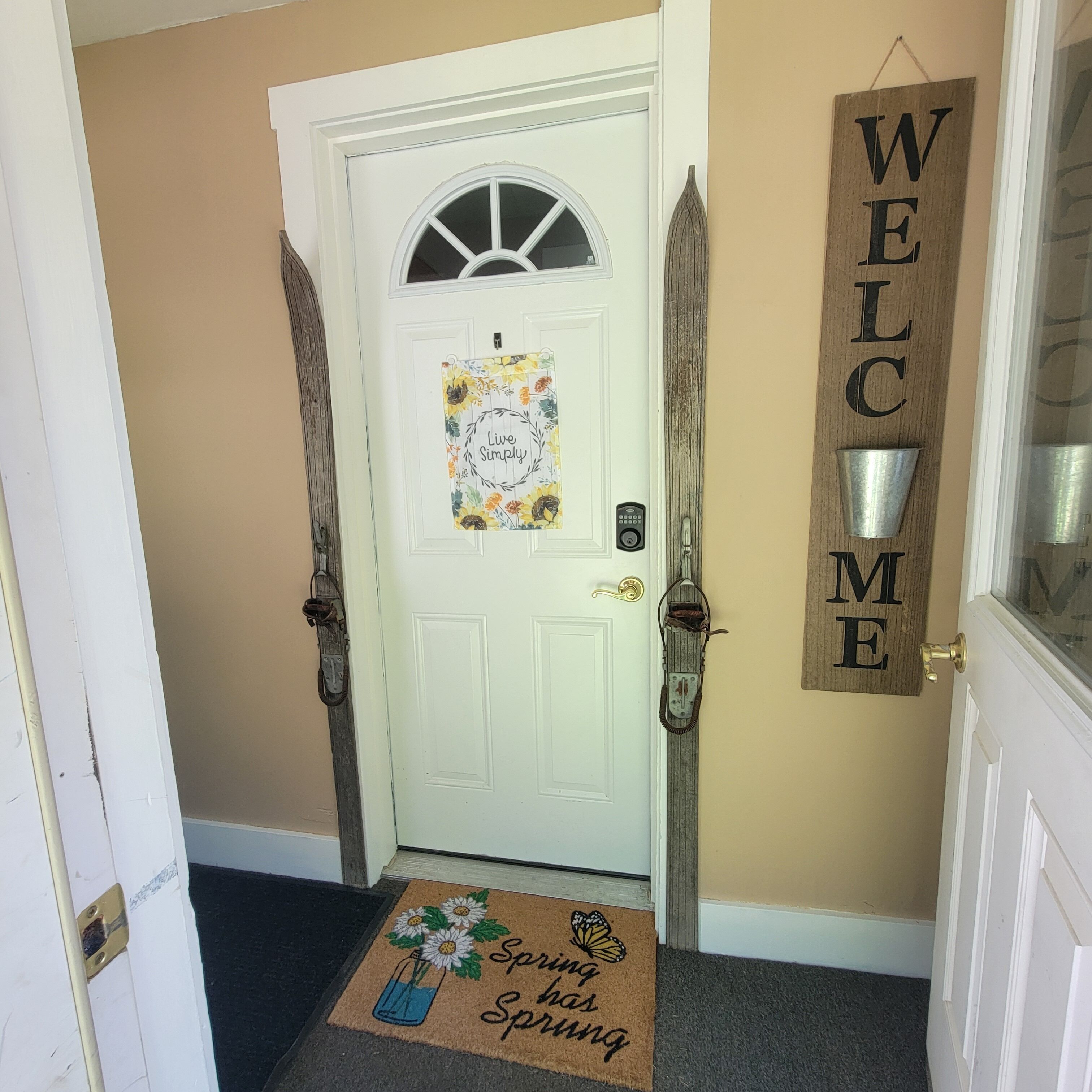A white door with a 'Live Simply' sign, a 'Welcome' sign, and a doormat with 'Spring has sprung' text.