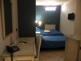 Una stanza con una scrivania, due letti, un comodino, una TV a parete e una finestra con tende.