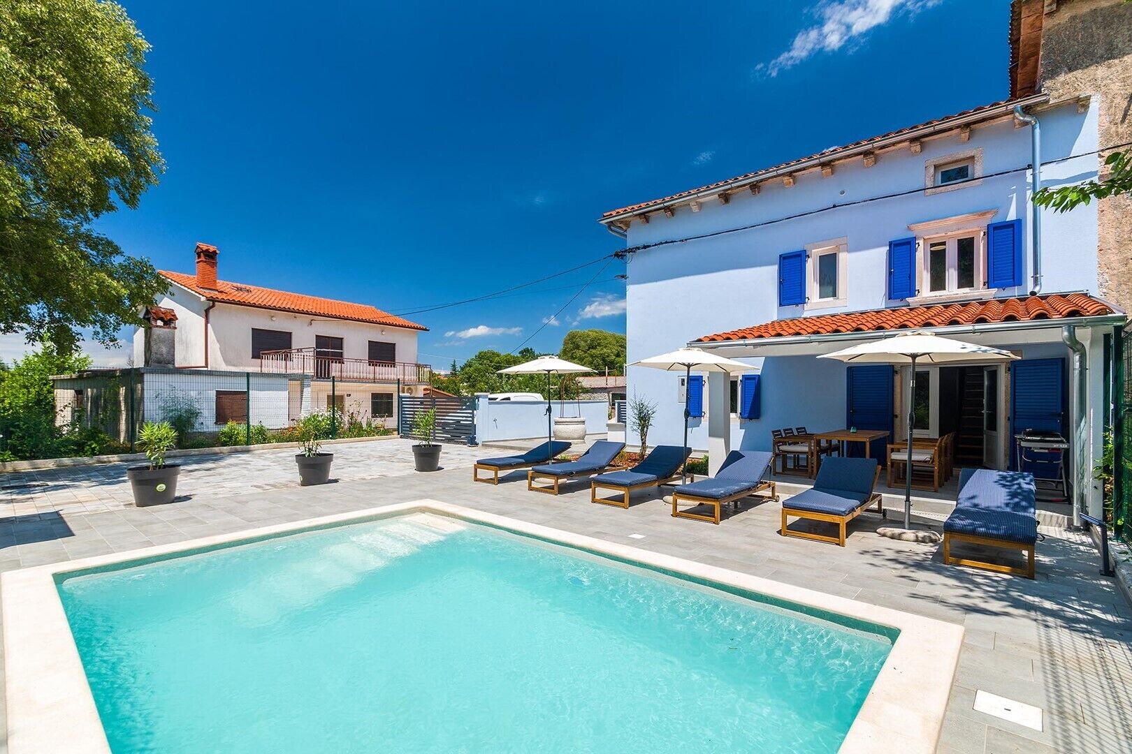 Une piscine entourée de bâtiments aux volets bleus, avec des chaises longues et des parasols.